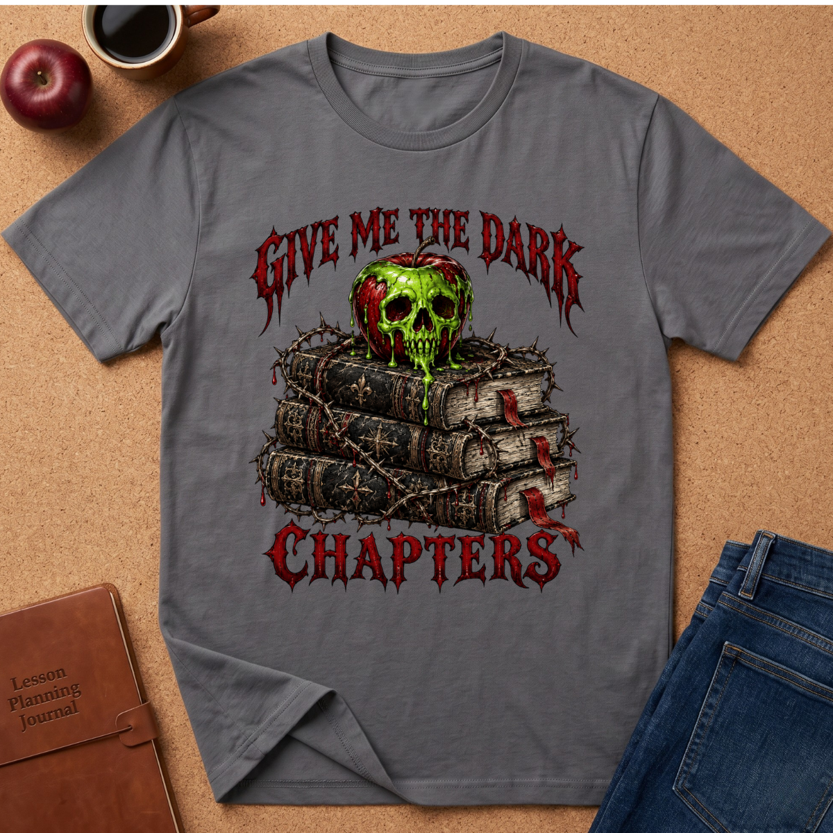 A gray short-sleeve graphic T-shirt laid flat on a cork surface, surrounded by lifestyle props including an apple, a coffee cup, a brown journal, and blue jeans. The shirt features a distressed red gothic-style text graphic reading 'GIVE ME THE DARK CHAPTERS' with a green-and-red skull resting on stacked books tied with barbed wire.