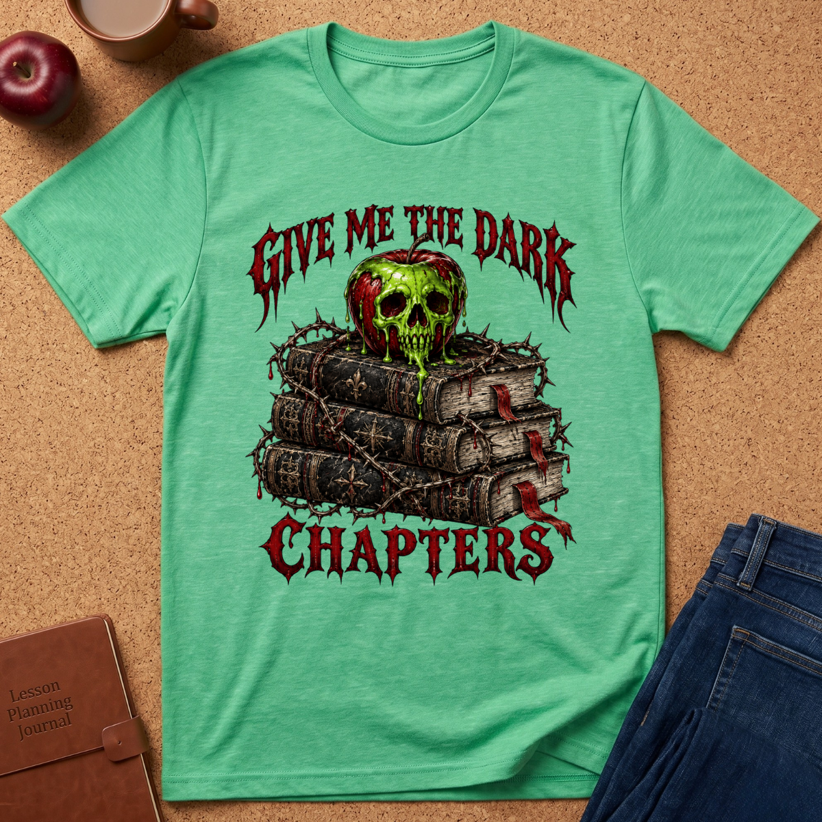 A mint green graphic t-shirt laid flat on a cork background, featuring a gothic design with the text 'GIVE ME THE DARK CHAPTERS' and an illustration of a green skull on top of stacked books bound with barbed wire. Styled flat-lay props include an apple, a coffee cup, a brown journal, and blue jeans.
