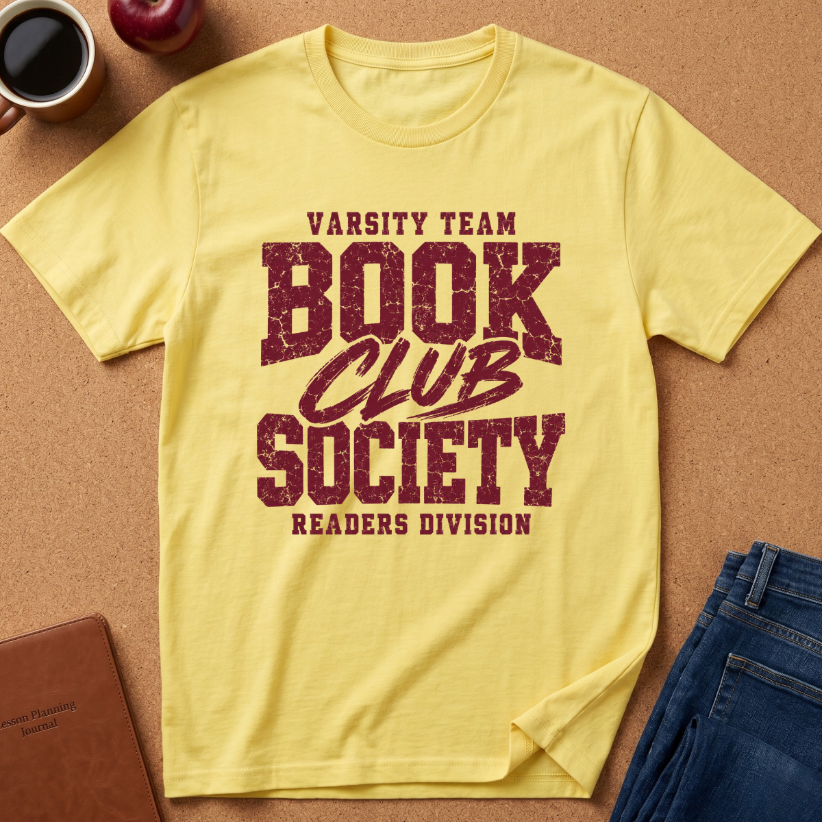 A pale yellow short-sleeve T-shirt laid flat on a cork-like surface, printed with distressed dark red text reading 'Varsity Team Book Club Society Readers Division.' Surrounding props include a coffee cup, apple, notebook, and folded blue jeans.