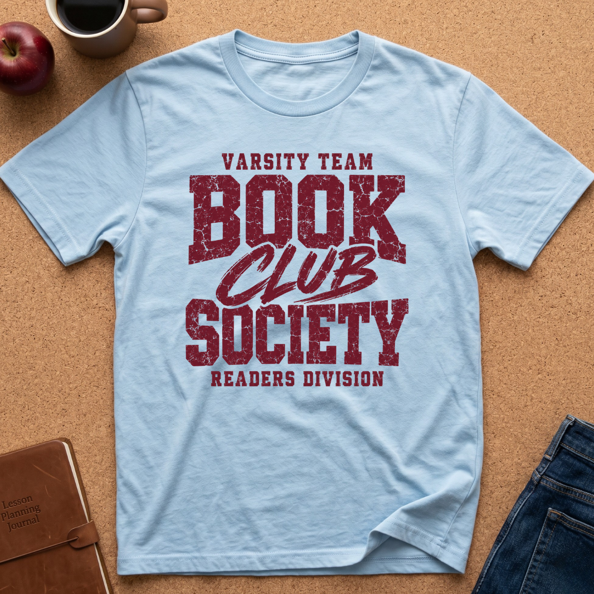A light blue crewneck t-shirt with a distressed dark red graphic reading 'VARSITY TEAM BOOK CLUB SOCIETY READERS DIVISION', laid flat on a cork background with a coffee mug, apple, notebook, and jeans as props.