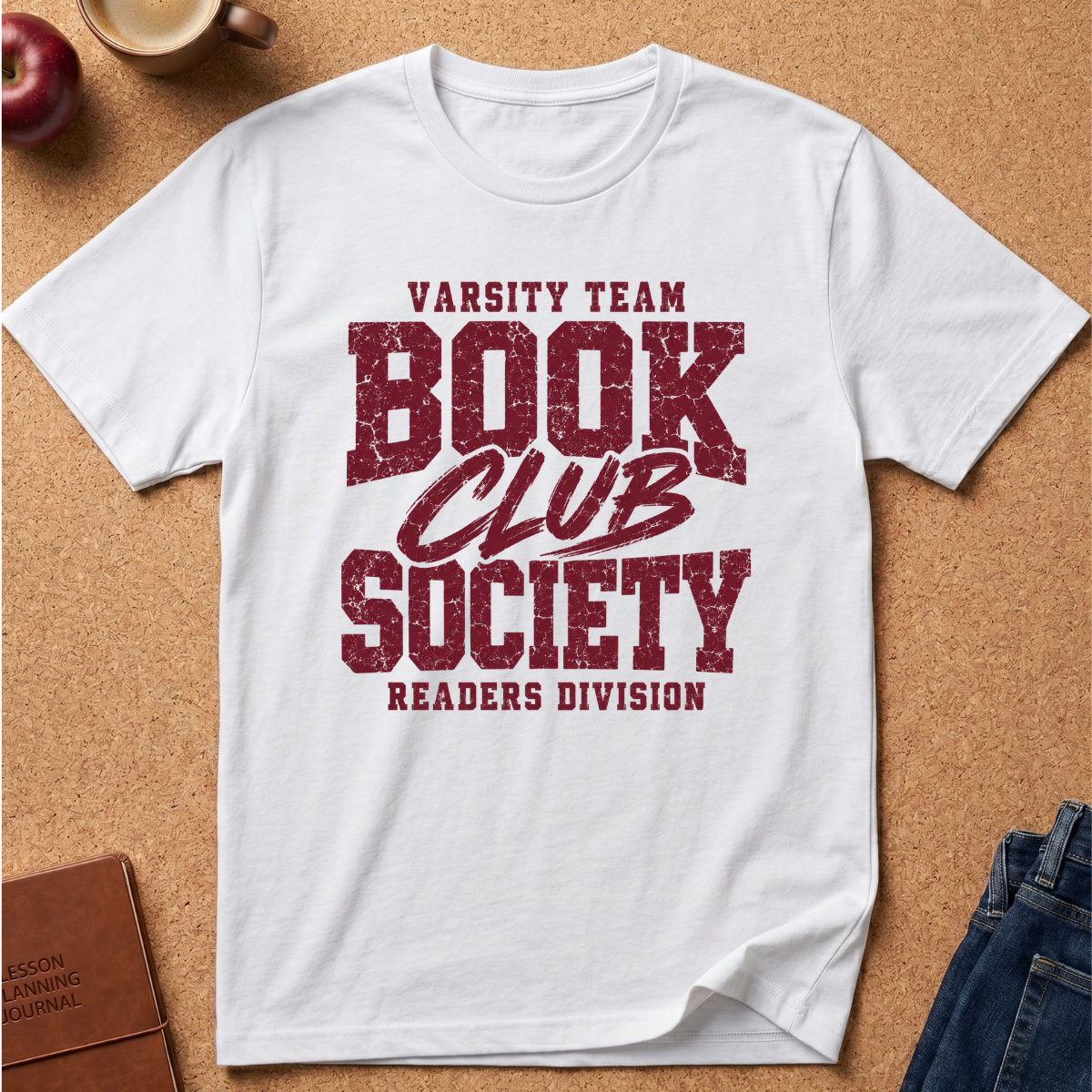 A white short-sleeve crewneck T-shirt laid flat on a cork background, featuring a distressed dark red graphic that reads 'VARSITY TEAM BOOK CLUB SOCIETY READERS DIVISION.' Styled flat-lay props include a book, apple, coffee cup, and jeans around the shirt.