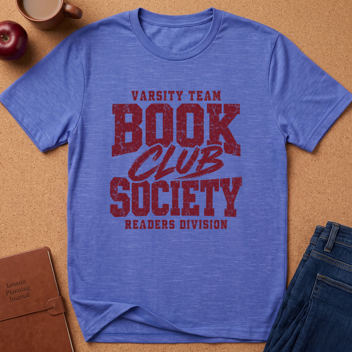 A heather blue short-sleeve t-shirt with a distressed red graphic reading 'Varsity Team Book Club Society Readers Division,' styled flat on a tan cork-like surface with a coffee mug, apple, notebook, and jeans around it.