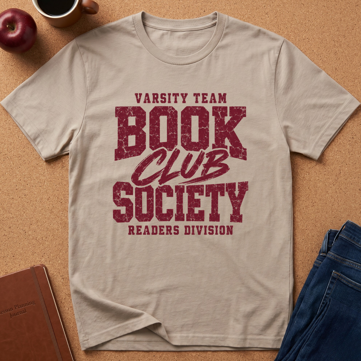 A beige graphic t-shirt laid flat on a cork background, featuring a distressed dark red print that reads 'VARSITY TEAM BOOK CLUB SOCIETY READERS DIVISION'. The shirt is styled with a coffee mug, an apple, a notebook, and blue jeans.