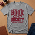 A heather gray short-sleeve t-shirt with a distressed red graphic reading 'VARSITY TEAM BOOK CLUB SOCIETY READERS DIVISION', laid flat on a cork background with an apple, coffee mug, notebook, and jeans around it.