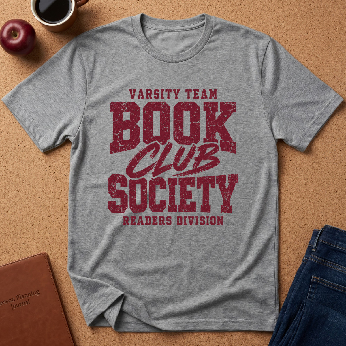 A heather gray short-sleeve t-shirt with a distressed red graphic reading 'VARSITY TEAM BOOK CLUB SOCIETY READERS DIVISION', laid flat on a cork background with an apple, coffee mug, notebook, and jeans around it.