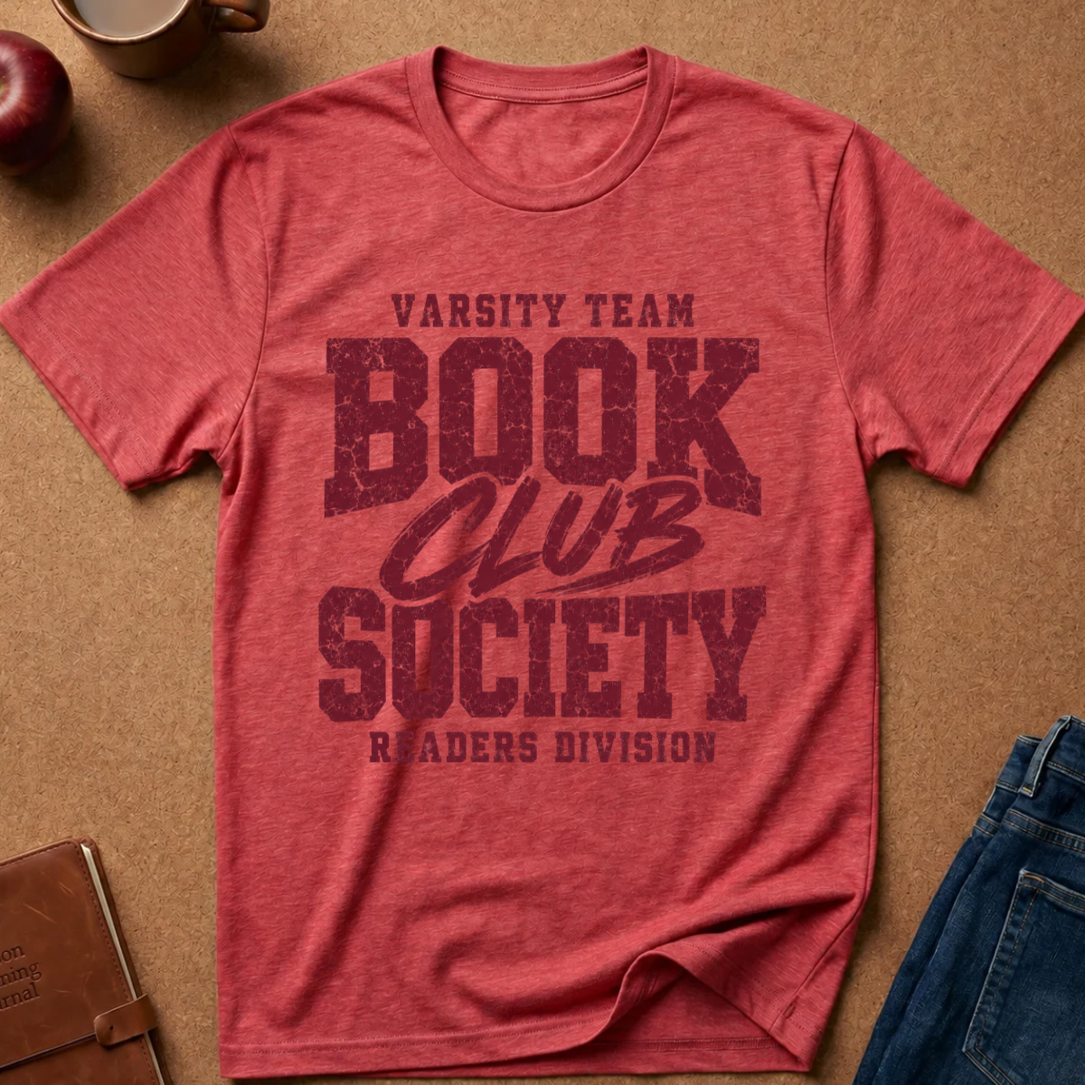 A heathered red short-sleeve t-shirt with a distressed dark red graphic reading 'VARSITY TEAM BOOK CLUB SOCIETY READERS DIVISION' laid flat on a brown kraft-paper background with partial jeans, an apple, and a notebook visible around it.