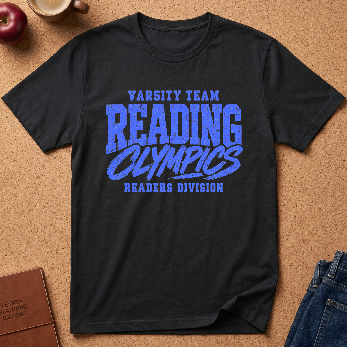A black short-sleeve graphic T-shirt laid flat on a cork-like surface, with blue text reading 'Varsity Team Reading Olympics Readers Division.' Surrounding props include part of a brown journal, a blue denim pocket, and a red apple.