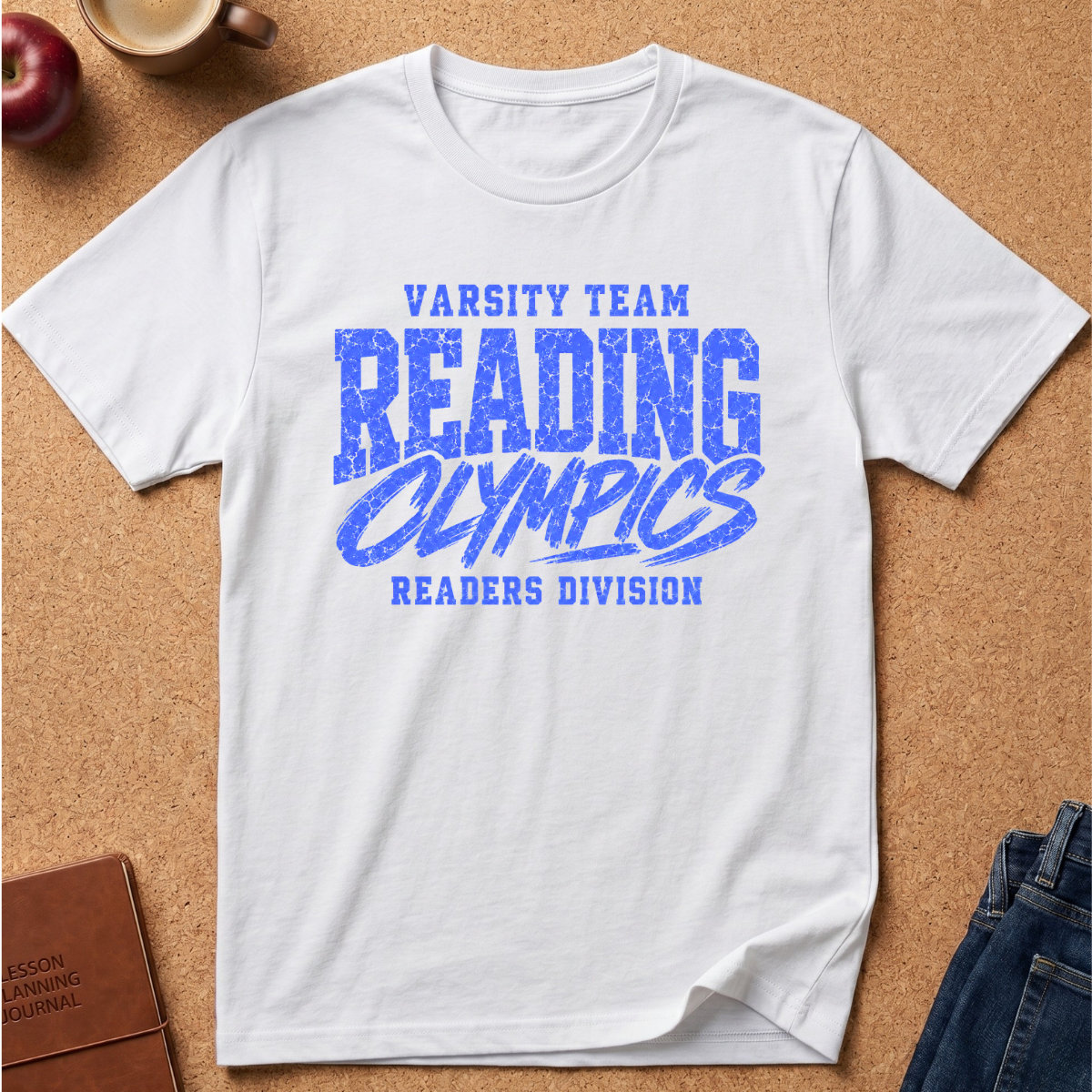 A white short-sleeve crewneck t-shirt with a blue distressed graphic reading 'VARSITY TEAM READING OLYMPICS READERS DIVISION', laid flat on a cork background with small lifestyle props including an apple, a mug, a journal, and jeans.