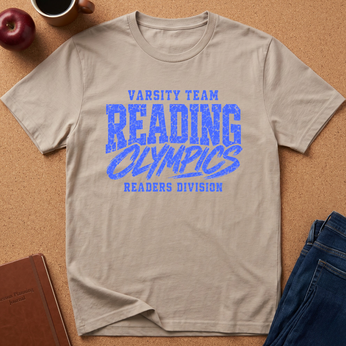 A beige crewneck t-shirt with a blue graphic reading 'VARSITY TEAM READING OLYMPICS READERS DIVISION' laid flat on a cork background with lifestyle props including an apple, coffee cup, notebook, and jeans.
