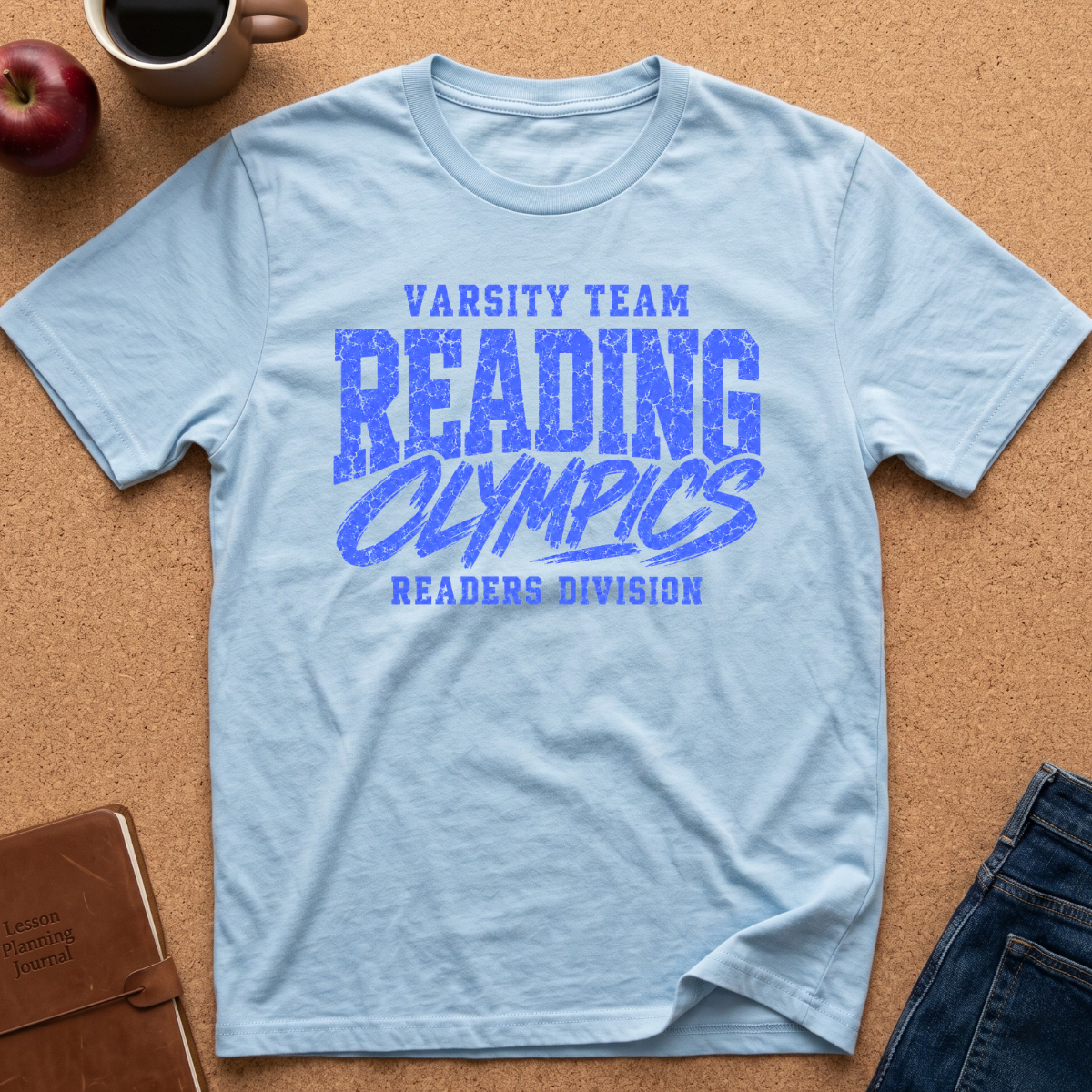 A light blue graphic t-shirt laid flat on a cork background, featuring the text 'VARSITY TEAM READING OLYMPICS READERS DIVISION' in blue. Styled flat-lay props include an apple, a coffee mug, a brown notebook, and blue jeans.