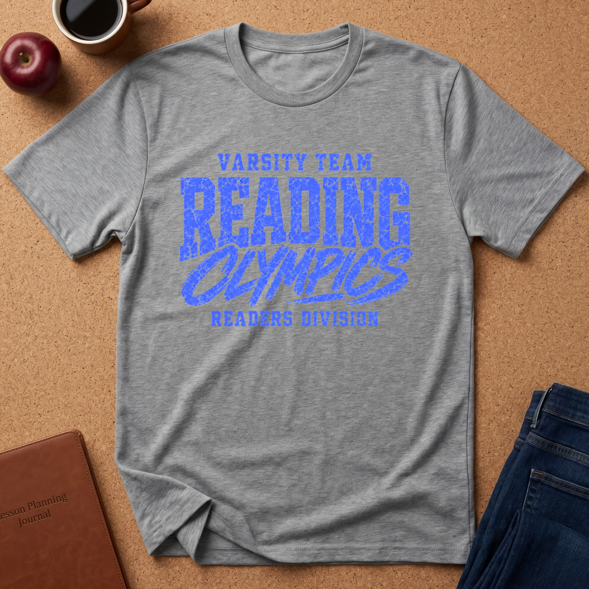 A heather gray short-sleeve t-shirt with a blue distressed graphic reading 'VARSITY TEAM READING OLYMPICS READERS DIVISION' laid flat on a cork background with a coffee cup, apple, notebook, and jeans as props.