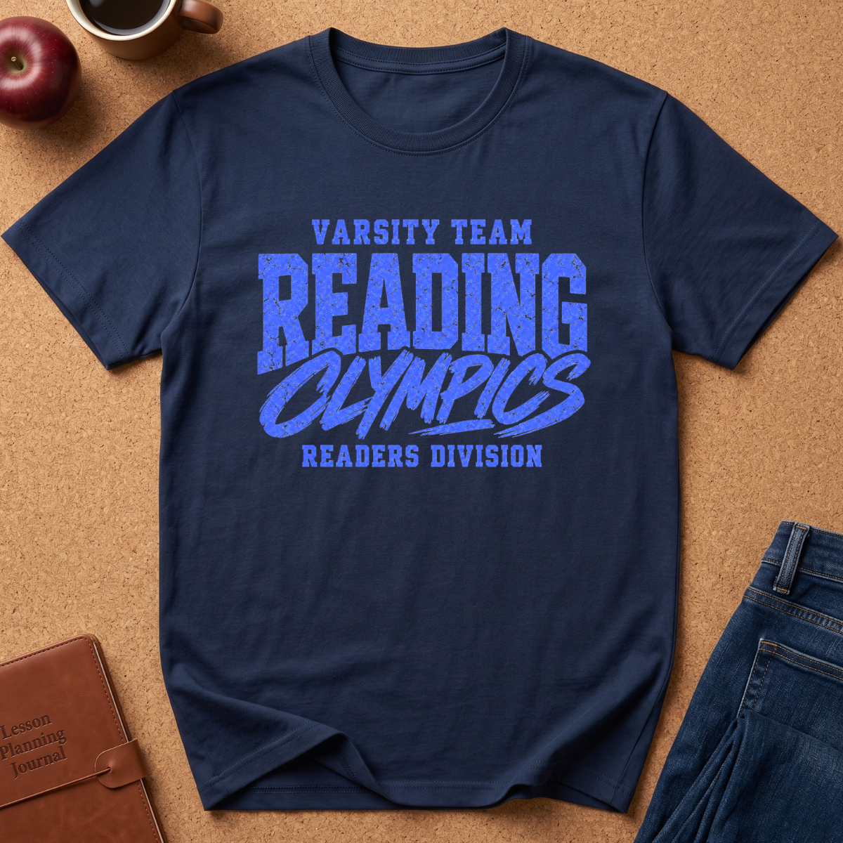 A navy blue short-sleeve t-shirt with a blue graphic reading 'VARSITY TEAM READING OLYMPICS READERS DIVISION' laid flat on a cork background with a red apple, coffee cup, leather journal, and jeans as props.