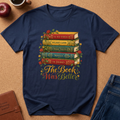 A navy blue short-sleeve t-shirt with a graphic of five colorful vintage books and the phrase 'The Book Was Better' in gold script, styled in a flat lay on a cork background with an apple, coffee cup, journal, and jeans nearby.