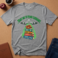 A heather gray graphic t-shirt laid flat on a cork background, featuring a green and multicolor design of a UFO dropping books into a stack labeled 'Library'. The shirt is styled with a coffee cup, apple, journal, and jeans.