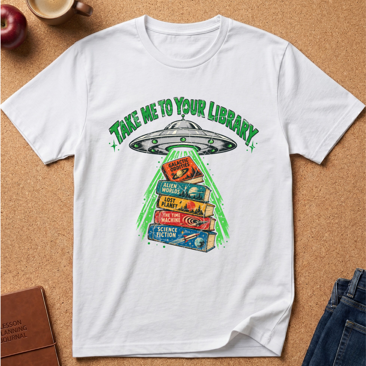 A white short-sleeve graphic T-shirt laid flat on a cork background, featuring a green and multicolor design of a UFO shining a beam of light onto a stack of science fiction books with the text 'TAKE ME TO YOUR LIBRARY'.