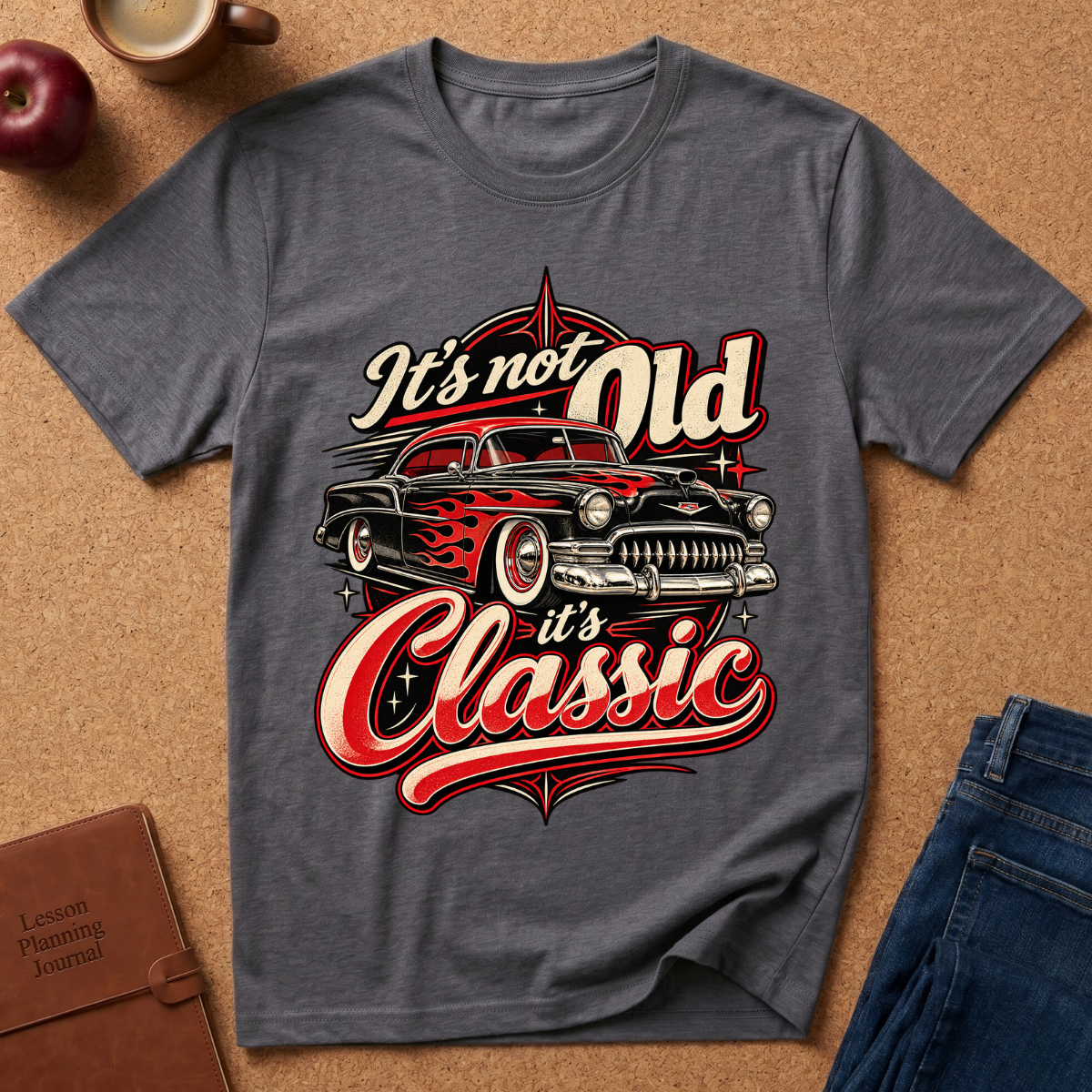 A dark gray graphic t-shirt laid flat on a brown cork background, featuring a vintage hot rod car and the text 'It's not Old it's Classic' in red and white. Styled flat-lay props include an apple, a coffee mug, a brown journal, and blue jeans.