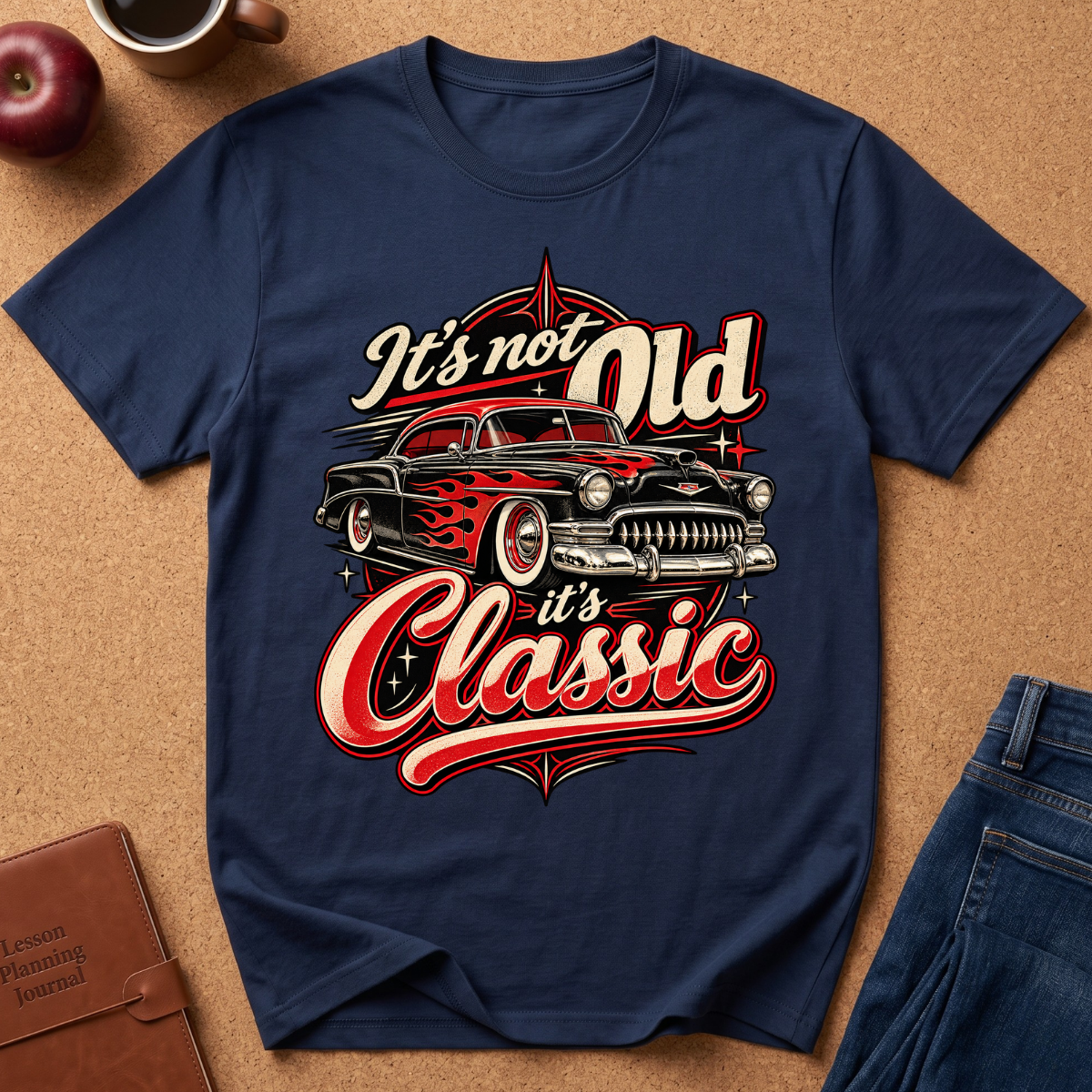 A navy blue graphic t-shirt laid flat on a cork background, featuring a retro-style illustration of a classic car with the text 'It's not Old it's Classic' in red, cream, and white. Styled flat-lay props include an apple, a coffee cup, a brown journal, and blue jeans.