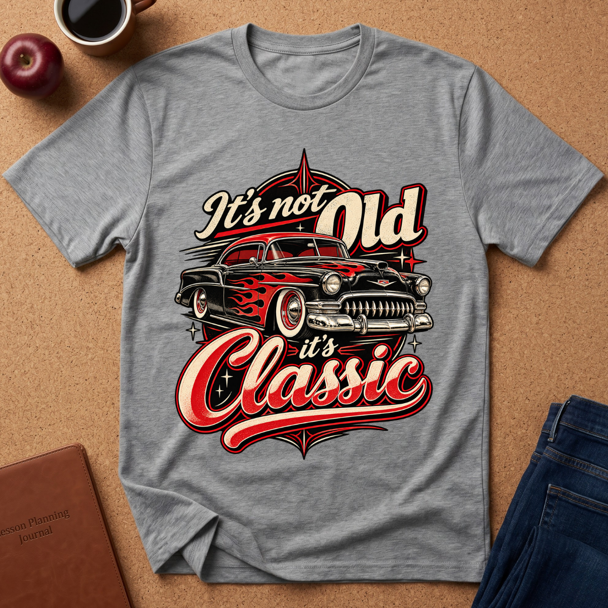 A heather gray graphic t-shirt laid flat on a cork background, featuring a vintage hot rod car design with the text 'It's not Old it's Classic'. The shirt is styled with an apple, a cup of coffee, a journal, and a pair of jeans.