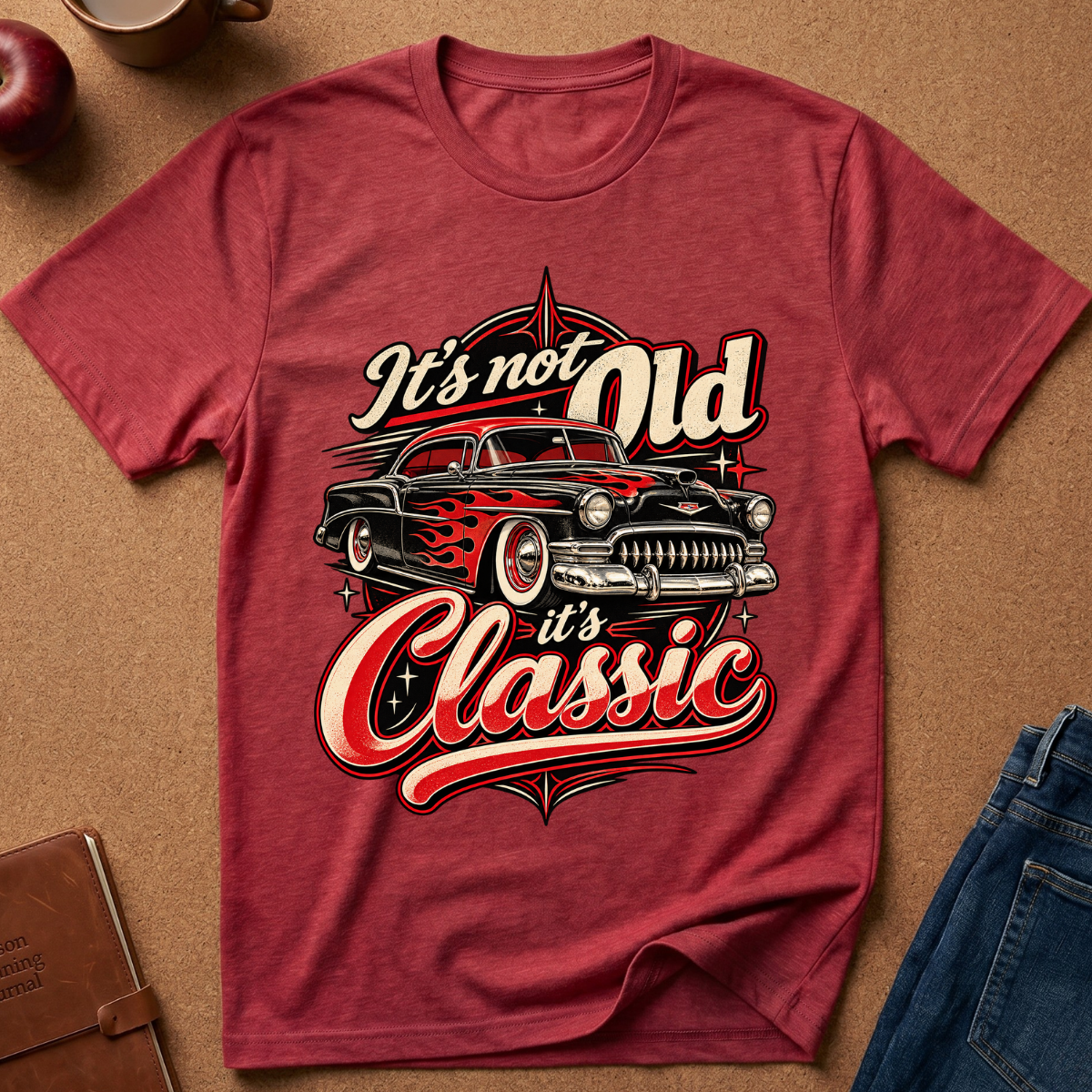 A heathered red graphic t-shirt laid flat on a brown textured background, featuring a vintage car and the text 'It's not Old it's Classic'.