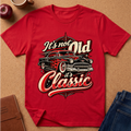 A red short-sleeve graphic T-shirt laid flat on a cork surface, featuring a vintage car print with the words “It’s not Old it’s Classic.” Styled flat-lay props include an apple, a mug, a notebook, and blue jeans around the shirt.