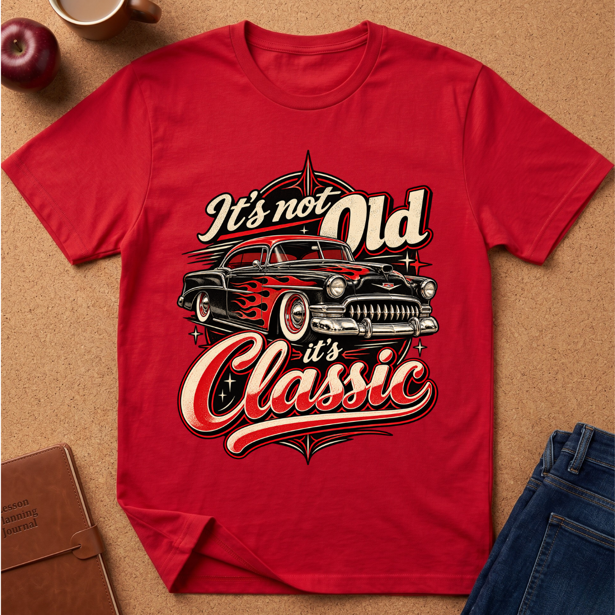 A red short-sleeve graphic T-shirt laid flat on a cork surface, featuring a vintage car print with the words “It’s not Old it’s Classic.” Styled flat-lay props include an apple, a mug, a notebook, and blue jeans around the shirt.