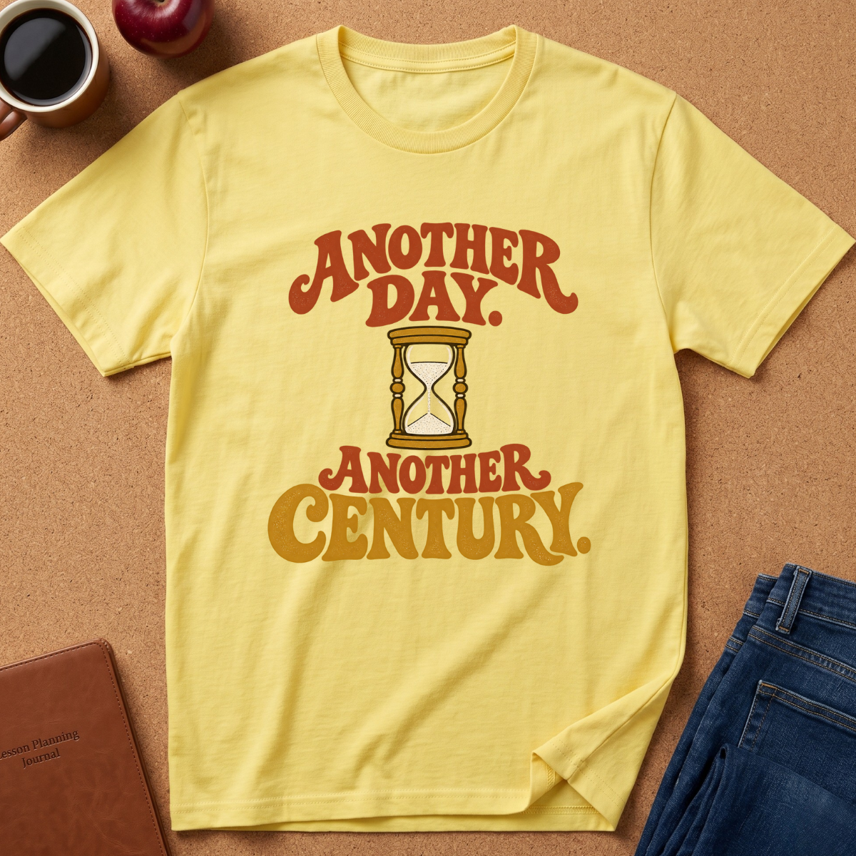 A pale yellow graphic T-shirt laid flat on a cork-like surface, with red and mustard text reading “ANOTHER DAY. ANOTHER CENTURY.