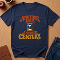 A navy blue graphic t-shirt laid flat on a cork background, featuring the text 'ANOTHER DAY. ANOTHER CENTURY.