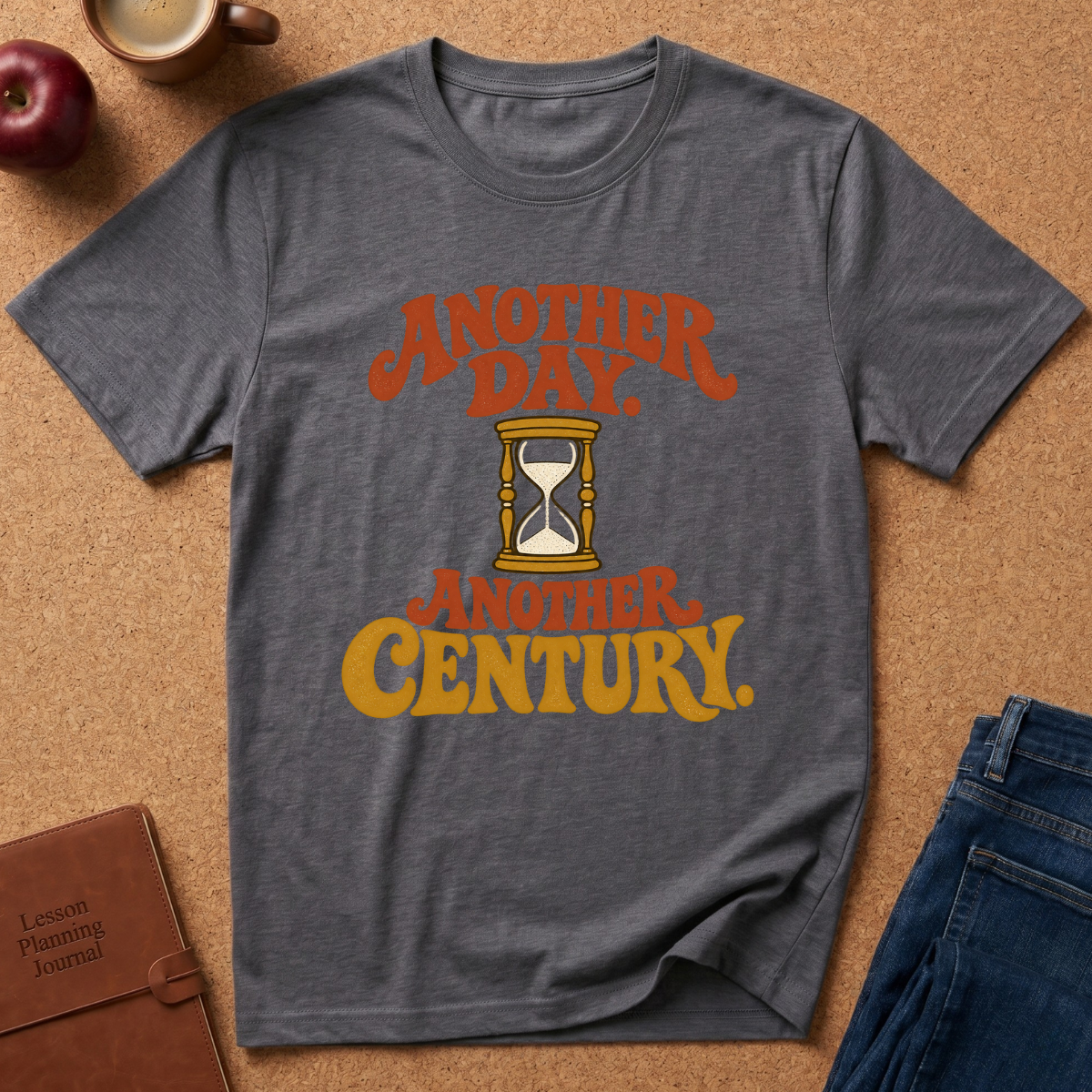 A dark heather gray graphic t-shirt laid flat on a cork background, featuring retro orange and yellow text that reads 'ANOTHER DAY. ANOTHER CENTURY.