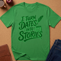 A bright green short-sleeve t-shirt with a dark green graphic reading 'I TURN DATES INTO STORIES' with a feather quill and calendar icon, laid flat on a cork surface with jeans, a mug, and an apple partially visible around it.