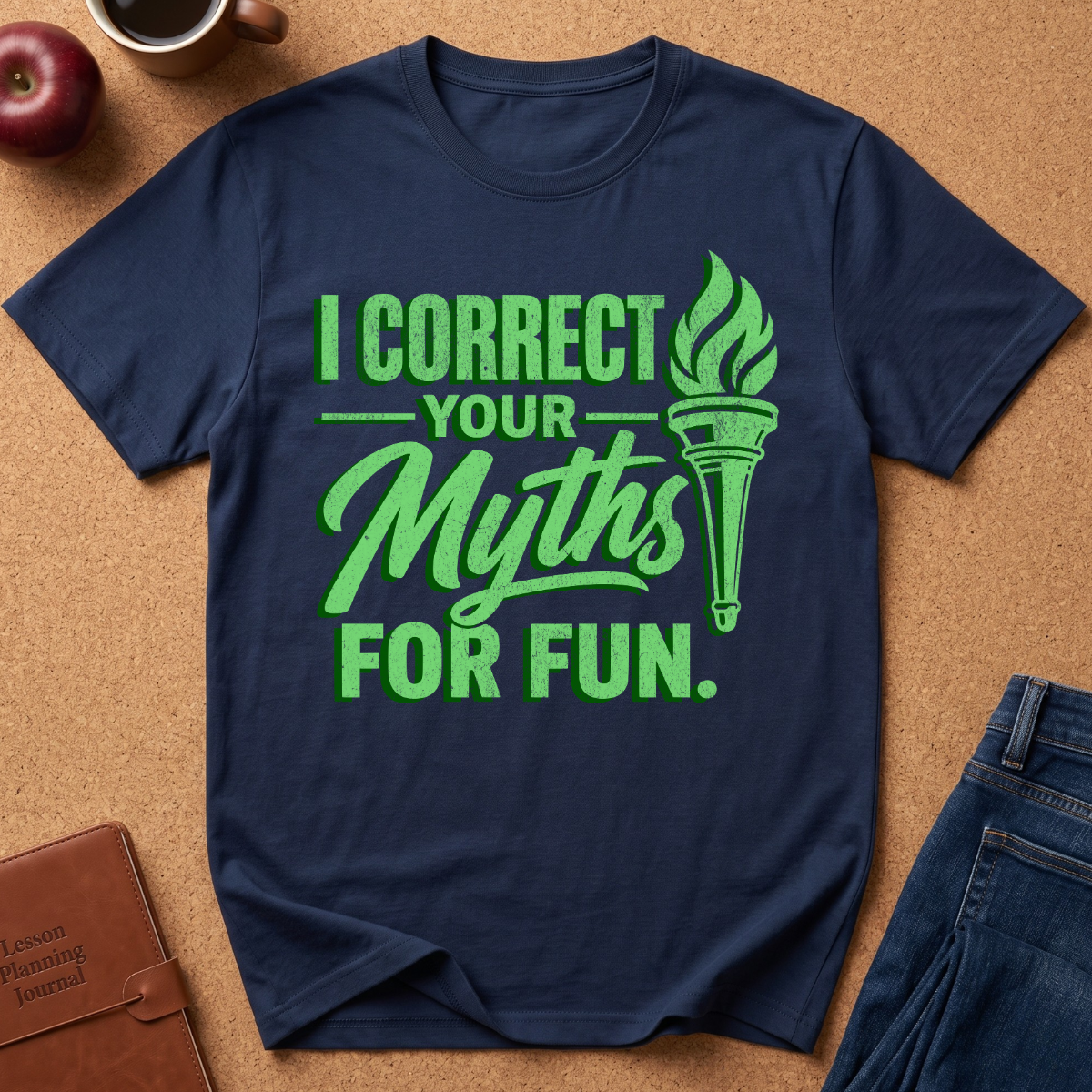 A navy blue short-sleeve t-shirt with a bright green graphic print that reads 'I CORRECT YOUR Myths FOR FUN.' alongside a torch illustration, laid flat on a brown cork background with partial props including an apple, a coffee cup, a notebook, and jeans.