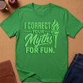 A green short-sleeve t-shirt laid flat on a cork background, featuring a light green graphic that reads 'I CORRECT YOUR MYTHS FOR FUN.' with a torch illustration.