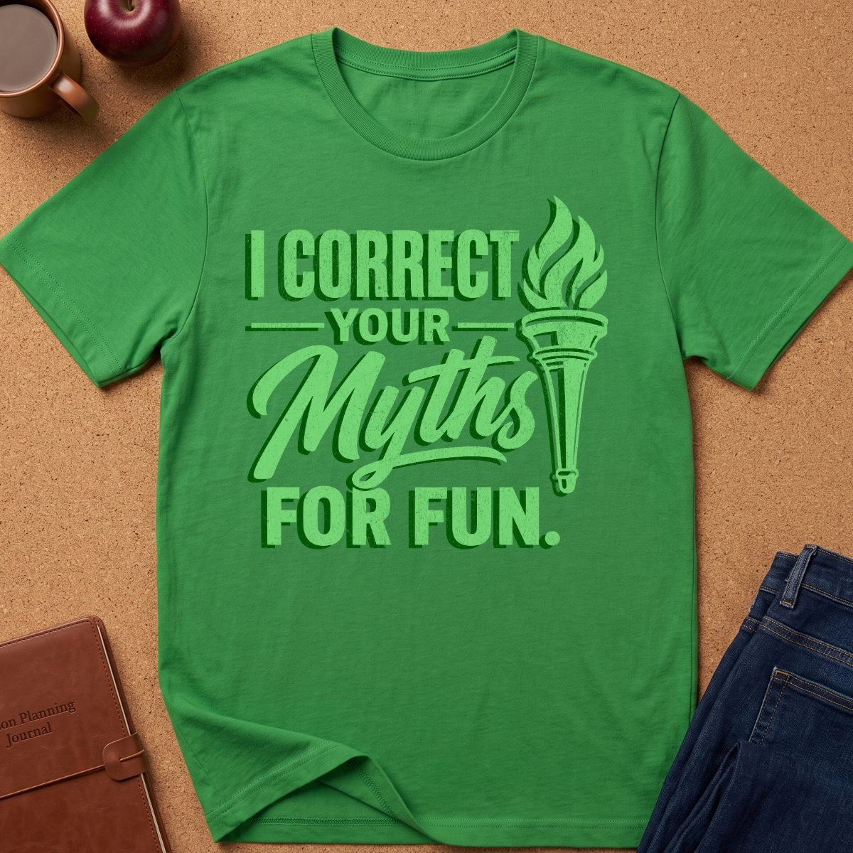 A green short-sleeve t-shirt laid flat on a cork background, featuring a light green graphic that reads 'I CORRECT YOUR MYTHS FOR FUN.' with a torch illustration.