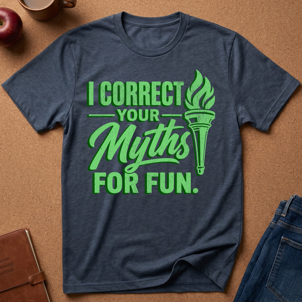 A navy blue heather graphic t-shirt laid flat on a brown cork background, featuring the text 'I CORRECT YOUR Myths FOR FUN.' in green next to a green torch graphic.