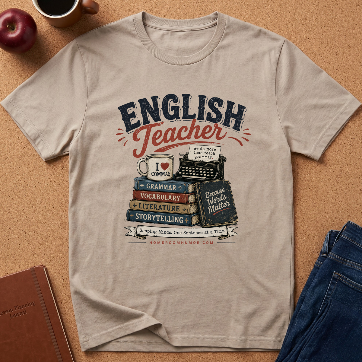 A beige graphic t-shirt laid flat on a cork background, featuring a retro-style 'English Teacher' design with books, a typewriter, a mug, and decorative text. Styled flat-lay props include an apple, a coffee cup, a notebook, and blue jeans.