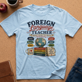 A light blue graphic t-shirt laid flat on a brown cork background, surrounded by lifestyle props including a coffee mug, an apple, a notebook, and jeans. The t-shirt features a colorful retro-style graphic that reads 'FOREIGN Language TEACHER' with illustrations of books, a globe, and language labels.