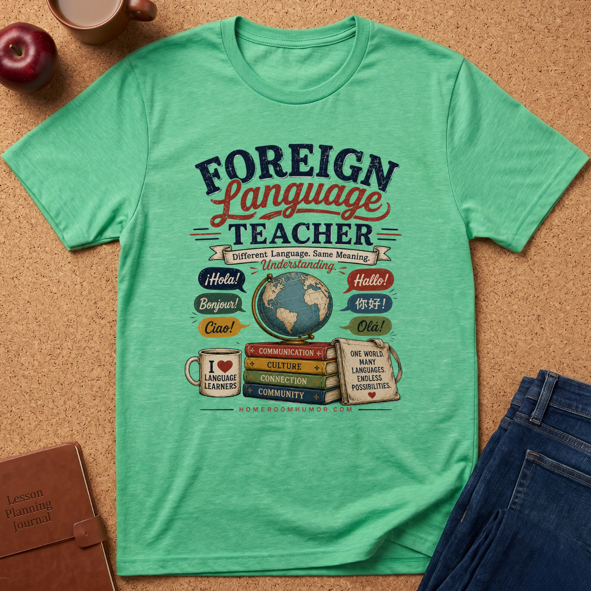 A mint green graphic t-shirt laid flat on a cork background, featuring a colorful 'Foreign Language Teacher' design with books, a globe, and greetings. Styled flat-lay props include an apple, coffee cup, brown journal, and blue jeans.