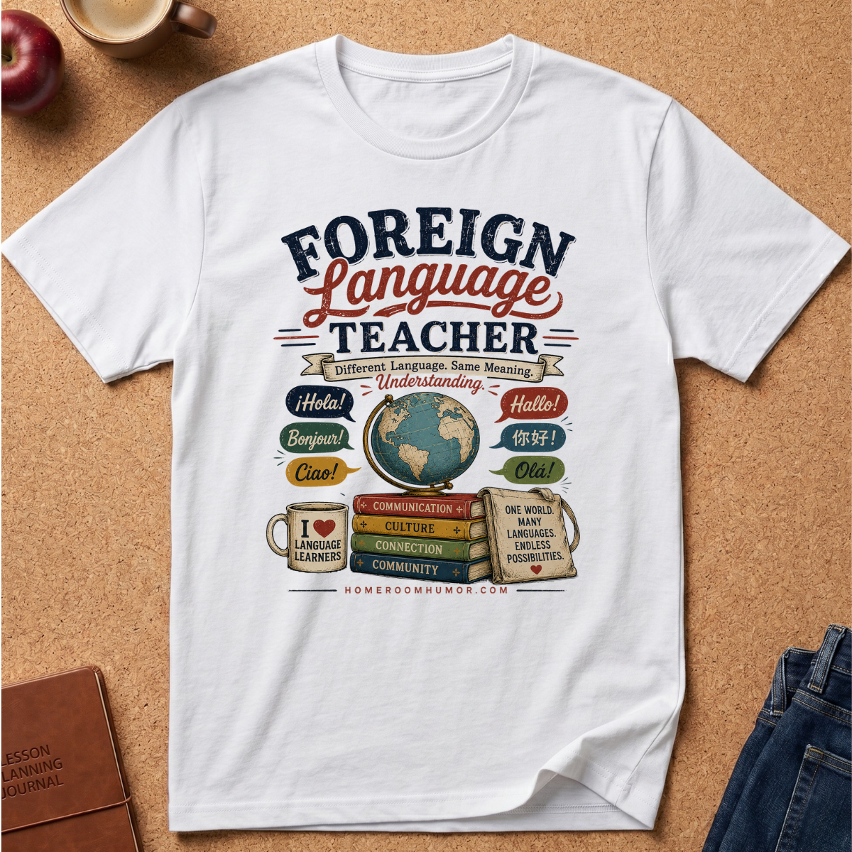 A white short-sleeve graphic T-shirt laid flat on a cork surface, featuring a colorful 'Foreign Language Teacher' design with books, a globe, and greeting bubbles. Partial props including an apple, coffee cup, notebook, and jeans are visible around the shirt.