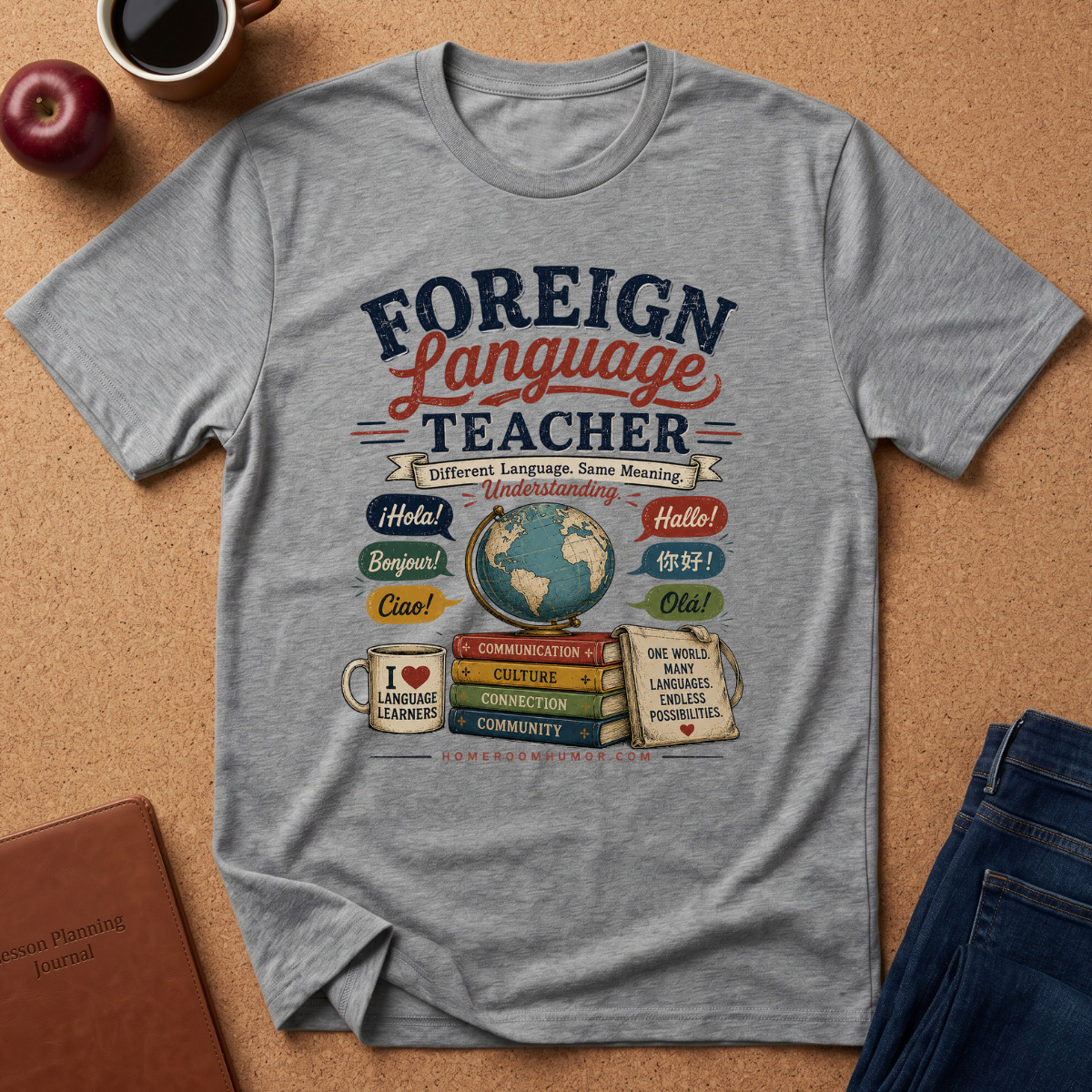 A heather gray graphic t-shirt laid flat on a cork background, featuring a colorful 'Foreign Language Teacher' design with a globe, books, and language flags. Styled flat-lay props include an apple, a coffee cup, a notebook, and blue jeans.