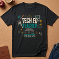 A black graphic t-shirt laid flat on a cork background, featuring a colorful 'Tech Ed Teacher' design with tools, laptops, and educational icons. Styled flat-lay props include an apple, a coffee cup, a journal, and jeans.