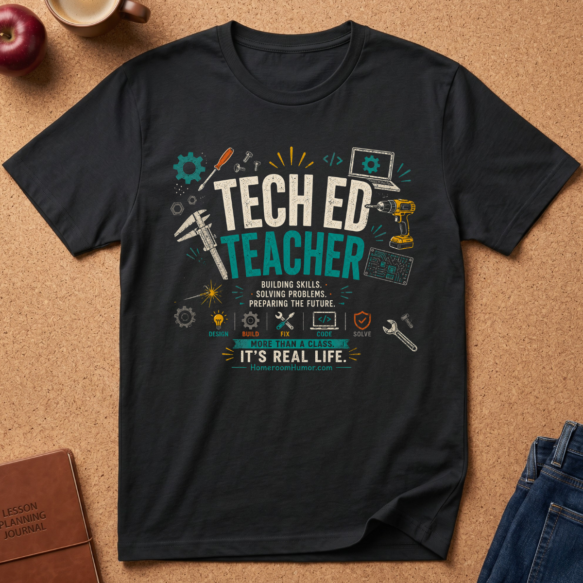 A black graphic t-shirt laid flat on a cork background, featuring a colorful 'Tech Ed Teacher' design with tools, laptops, and educational icons. Styled flat-lay props include an apple, a coffee cup, a journal, and jeans.