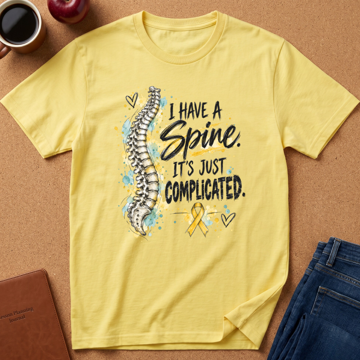 A yellow graphic t-shirt laid flat on a cork background, featuring a spine illustration and the text 'I HAVE A Spine. IT'S JUST COMPLICATED.