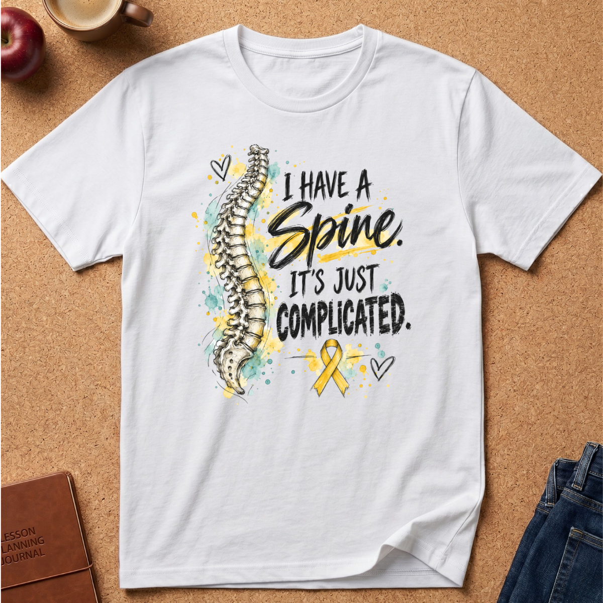 A white short-sleeve graphic T-shirt laid flat on a cork background, featuring a spine illustration and the text 'I HAVE A Spine. IT'S JUST COMPLICATED.