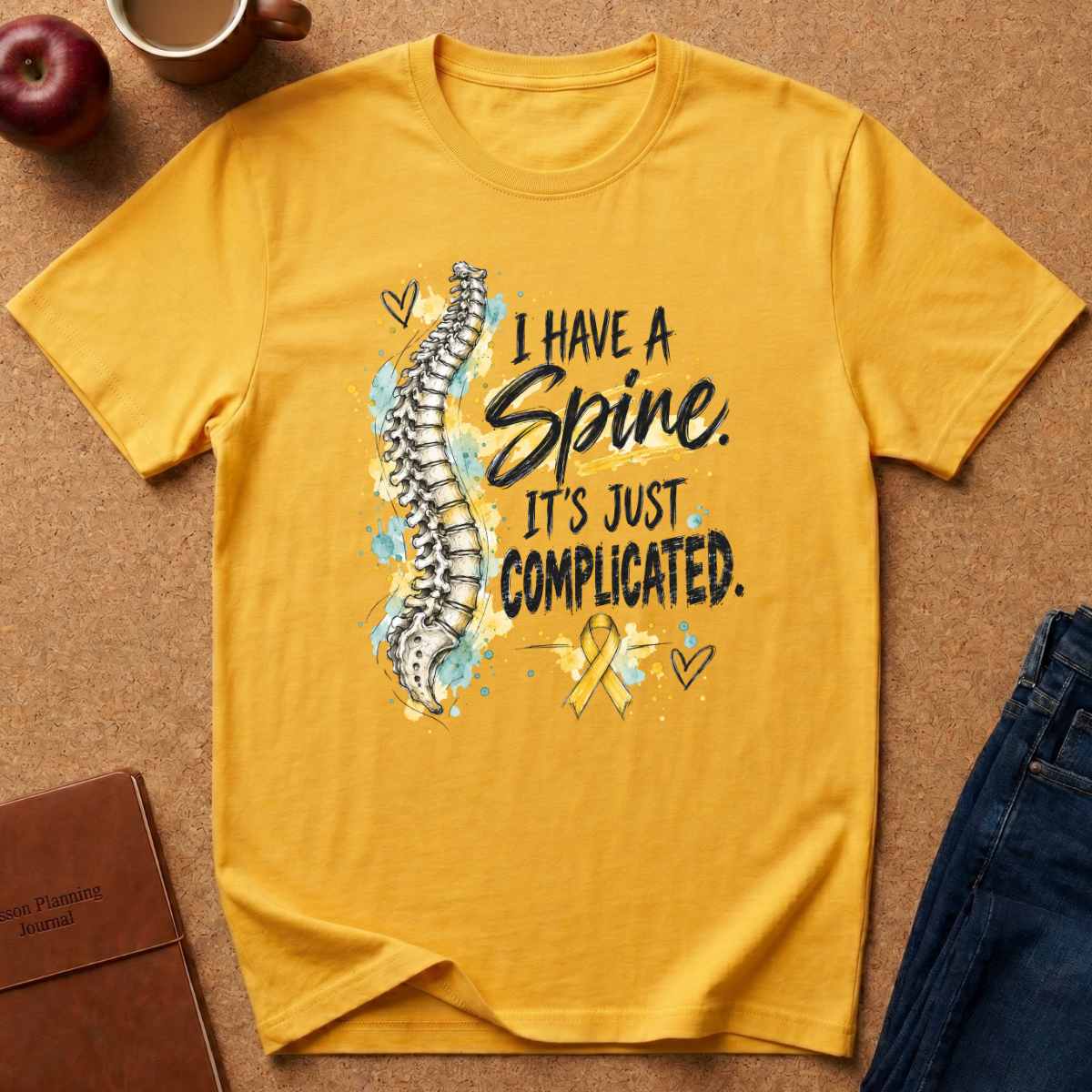A yellow graphic t-shirt laid flat on a brown textured surface, featuring a watercolor spine illustration and the text 'I HAVE A Spine. IT'S JUST COMPLICATED.