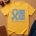 A yellow short-sleeve T-shirt laid flat on a cork surface, featuring a blue awareness ribbon made of puzzle pieces and the text 'THEIR FIGHT IS MY FIGHT' with 'Spina Bifida Awareness' below. Surrounding props include an apple, a mug, a notebook, and jeans.