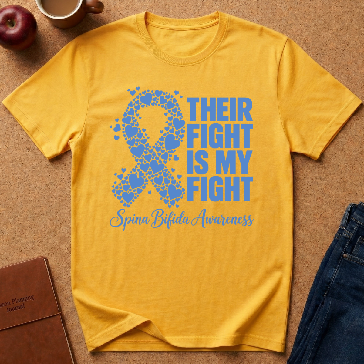 A yellow short-sleeve T-shirt laid flat on a cork surface, featuring a blue awareness ribbon made of puzzle pieces and the text 'THEIR FIGHT IS MY FIGHT' with 'Spina Bifida Awareness' below. Surrounding props include an apple, a mug, a notebook, and jeans.
