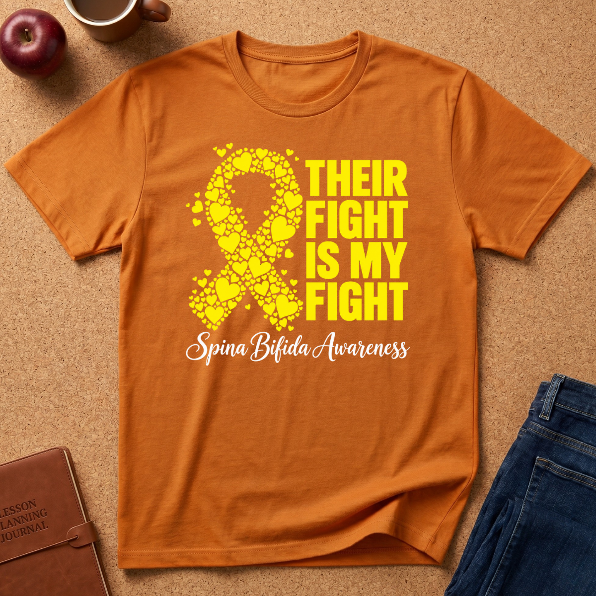 An orange short-sleeve t-shirt laid flat on a cork surface, featuring a yellow awareness ribbon made of shattered glass and the text 'THEIR FIGHT IS MY FIGHT' and 'Spina Bifida Awareness' in yellow and white script. A brown leather journal, a red apple, a mug, and blue jeans are styled around it.