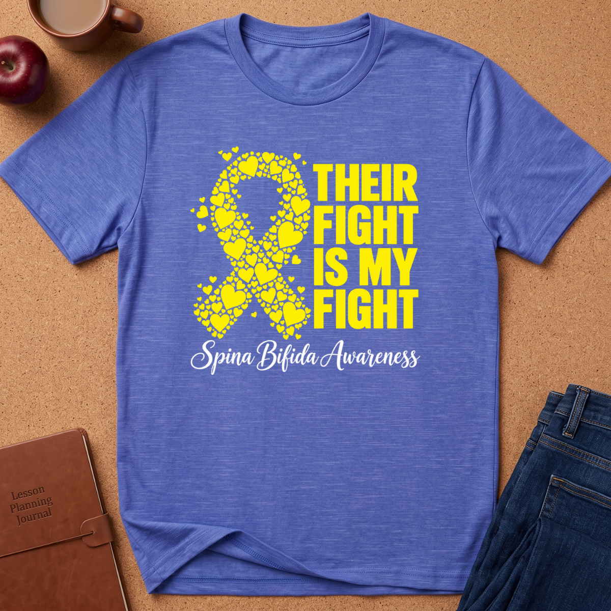 A heathered blue short-sleeve t-shirt laid flat on a cork surface, featuring a yellow ribbon made of small hearts and the text 'THEIR FIGHT IS MY FIGHT' with 'Spina Bifida Awareness' in white script below. A brown notebook, apple, coffee cup, and jeans are styled around it as props.
