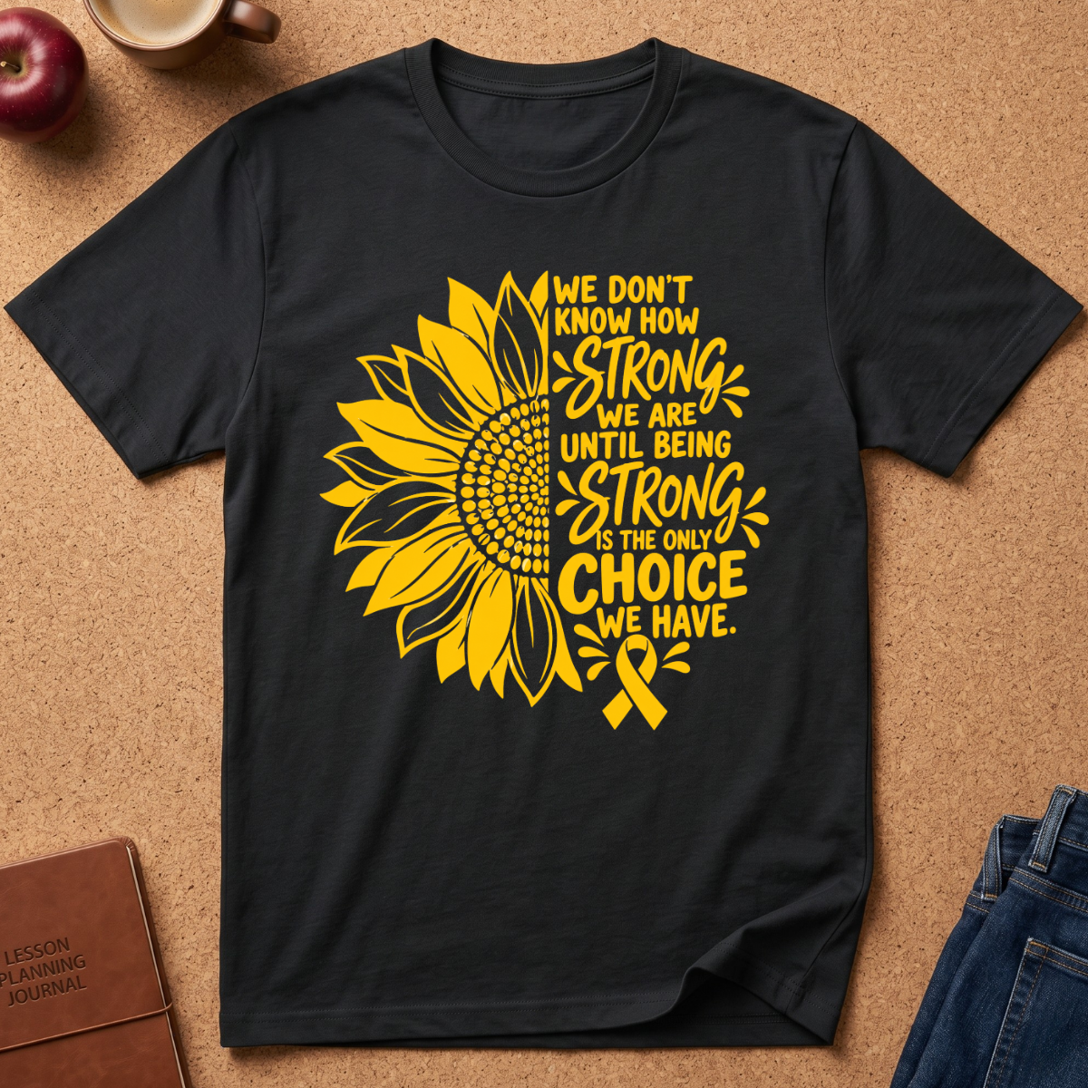 A black short-sleeve graphic T-shirt laid flat on a cork-like surface, featuring a large yellow sunflower and the text 'We don't know how strong we are until being strong is the only choice we have,' alongside a yellow awareness ribbon. Partial props around the shirt include an apple, a mug, a journal, and jeans.