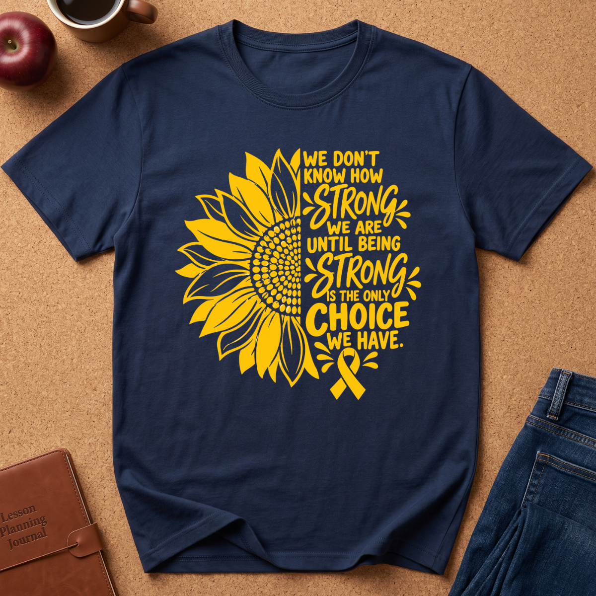 A navy blue short-sleeve t-shirt with a yellow sunflower graphic and the text 'WE DON'T KNOW HOW STRONG WE ARE UNTIL BEING STRONG IS THE ONLY CHOICE WE HAVE.' paired with a yellow ribbon, laid flat on a cork background with jeans, a book, and an apple nearby.