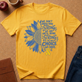 A yellow short-sleeve T-shirt laid flat on a brown cork-like surface, featuring a large blue sunflower graphic and the text 'We don't know how strong we are until being strong is the only choice we have,' plus a yellow ribbon. A red apple, coffee cup, notebook, and jeans are partially visible around it as styling props.
